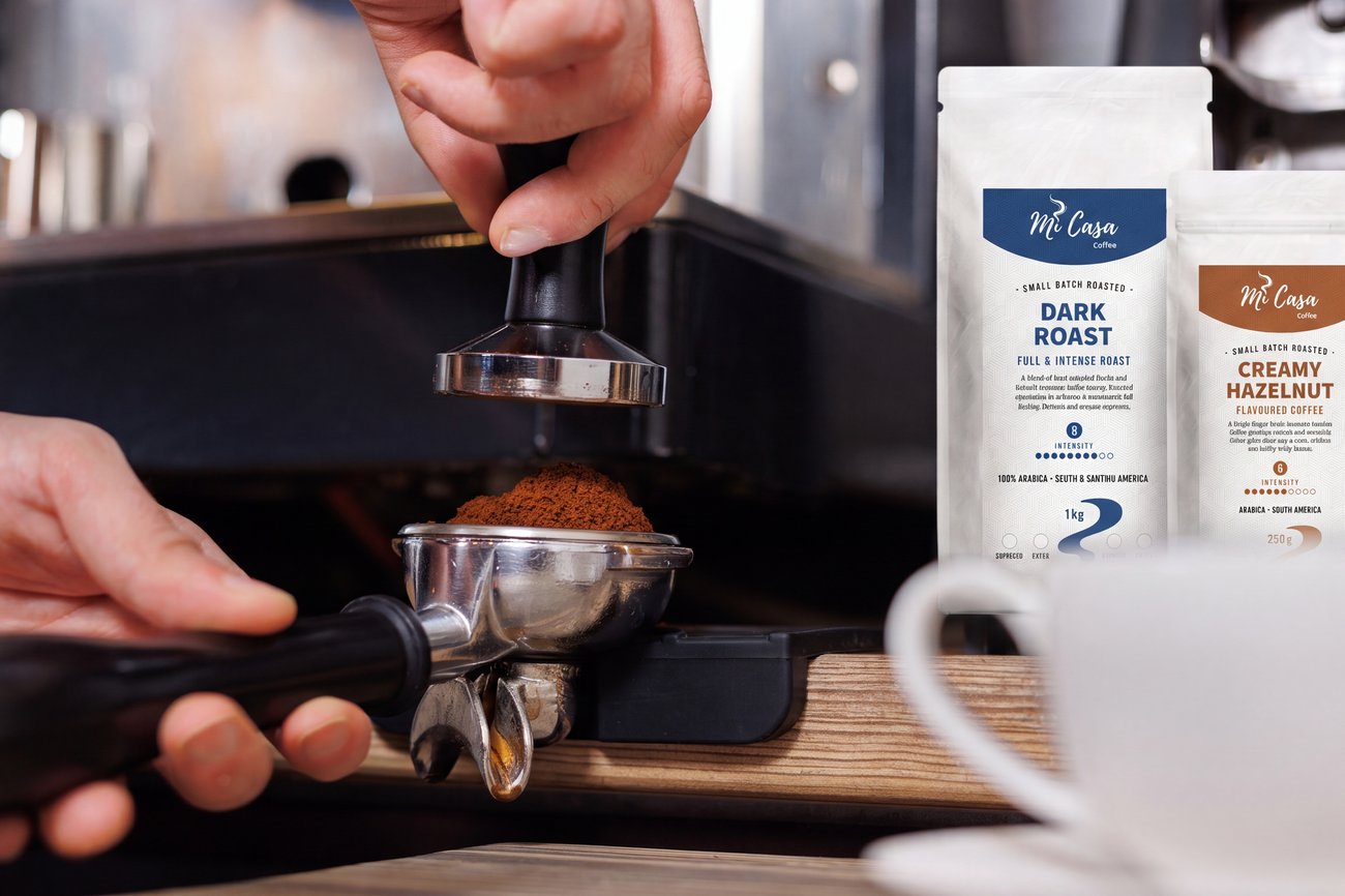A barista tamping fresh grounds beside MiCasa Dark Roast and Creamy Hazelnut bags