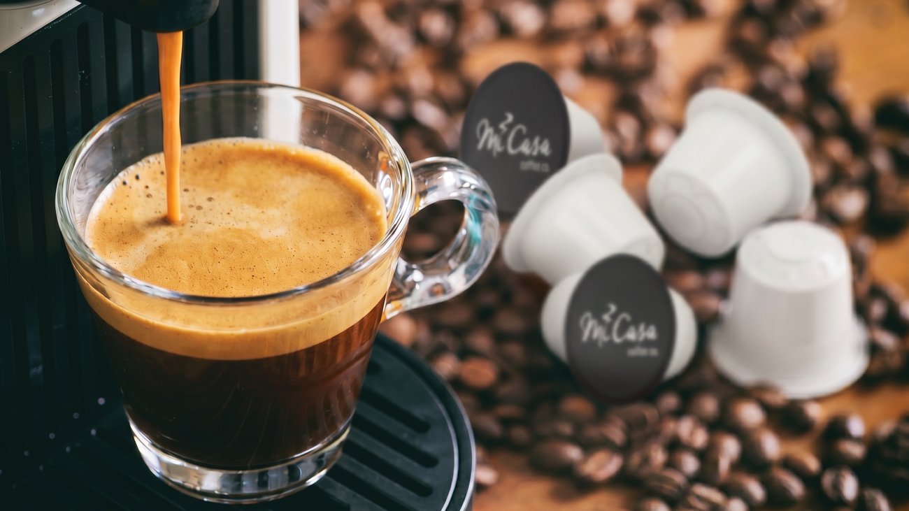 A rich espresso pulling into a glass cup beside MiCasa Nespresso-compatible capsules and freshly roasted beans