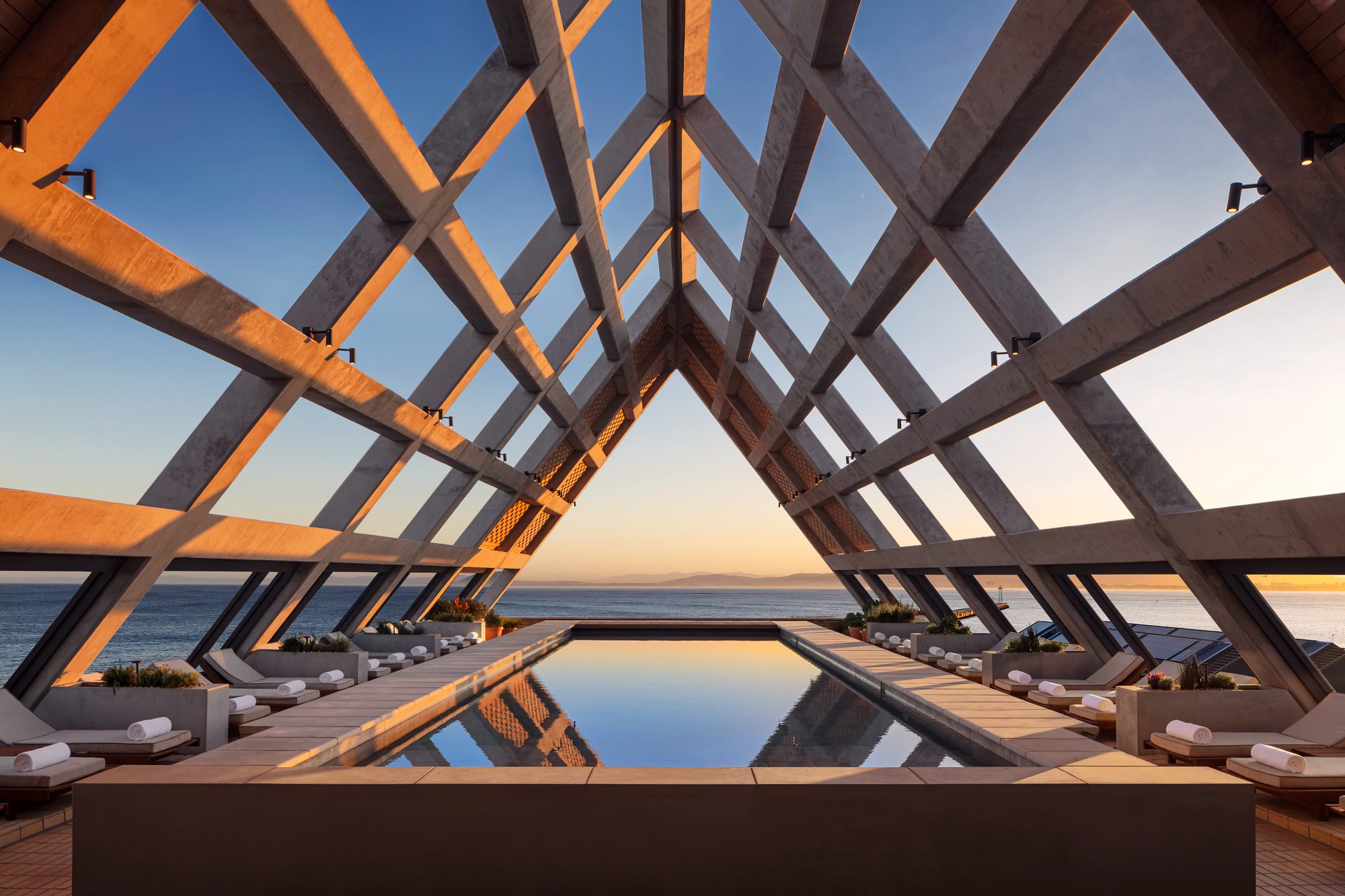 The rooftop pool of The Cape Town EDITION under a timber A-frame roof at sunset, with the Atlantic beyond