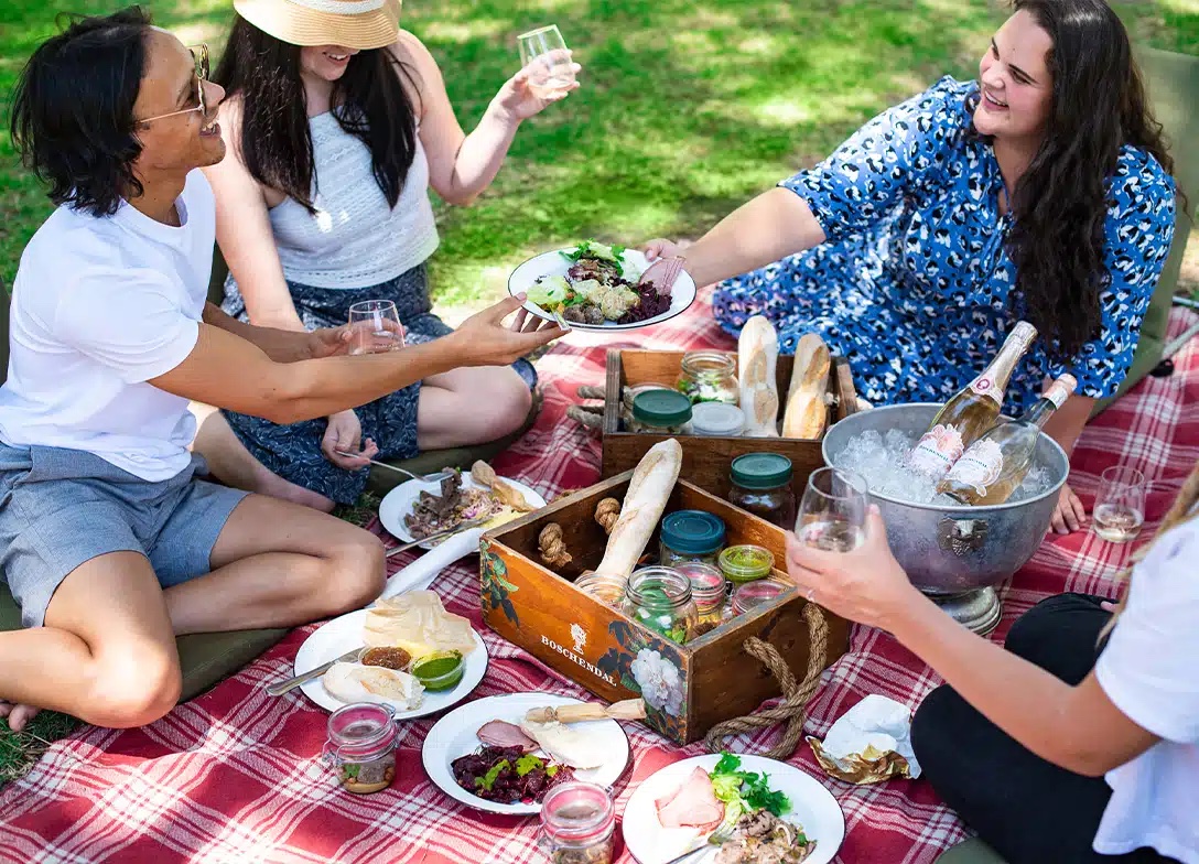 Friends share a picnic of salads, bread and wine on a red checked blanket on the lawn at Boschendal