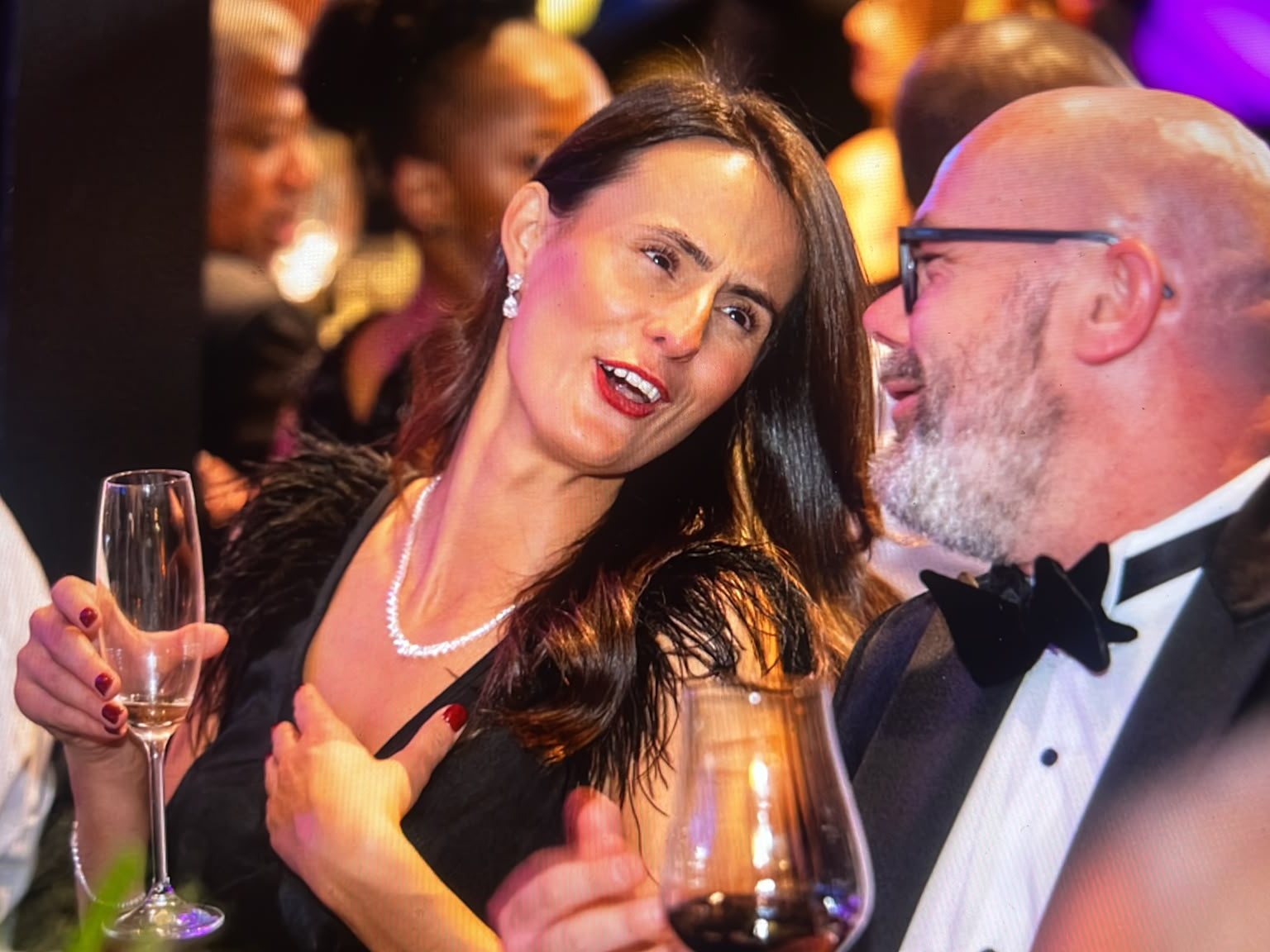 A guest in a black dress holding a glass of champagne talks with a bearded man in a dinner suit and velvet bow tie.
