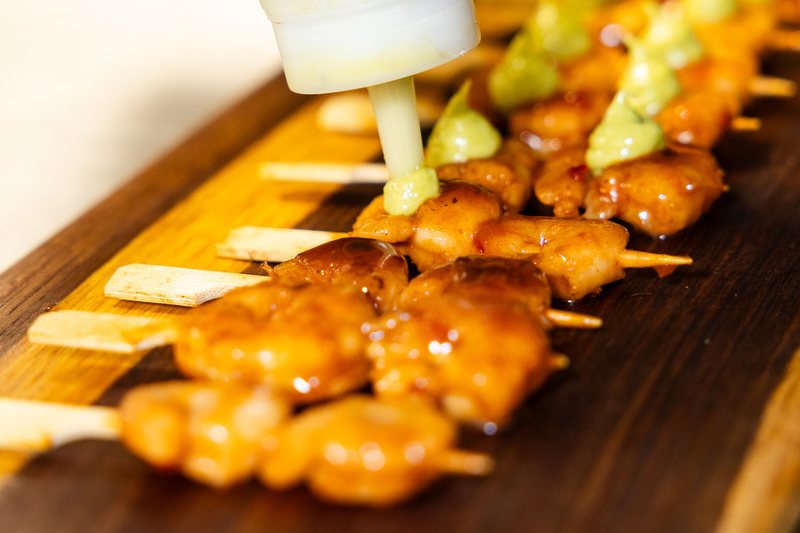 Glazed skewers lined up on a wooden board, each finished with a piped dot of green sauce.