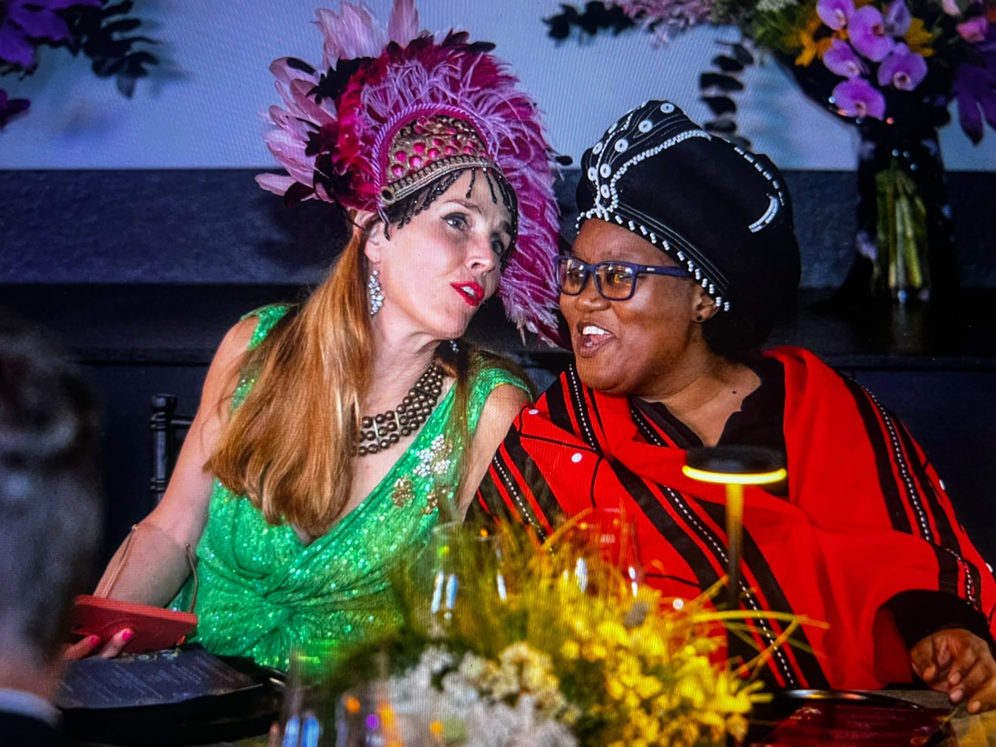 Two guests in conversation at a dinner table: one in a green sequinned dress and a pink feathered headdress, the other in a red and black blanket with a beaded black headwrap, with orchids behind.