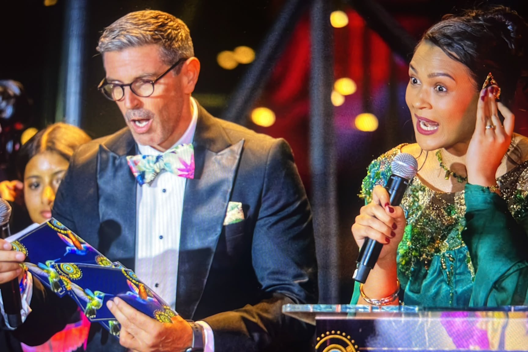 A man in a dinner suit and bow tie and a woman in an emerald gown speaking into microphones on stage, both looking surprised.