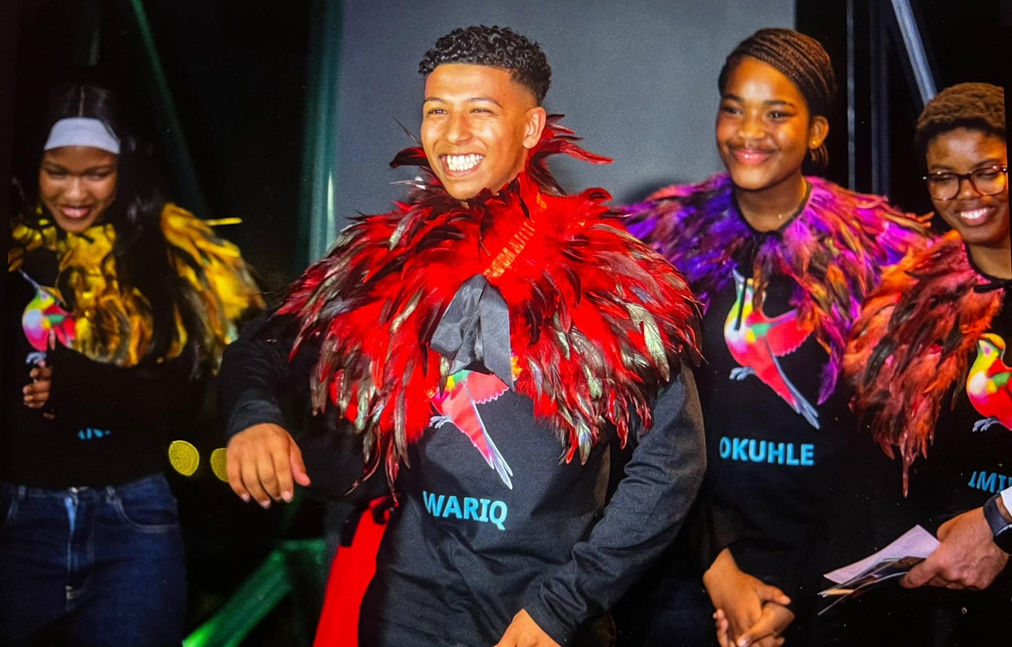 Young people in feathered capes of red, purple and green, worn over Hummingbird Ball T-shirts, smiling on stage.