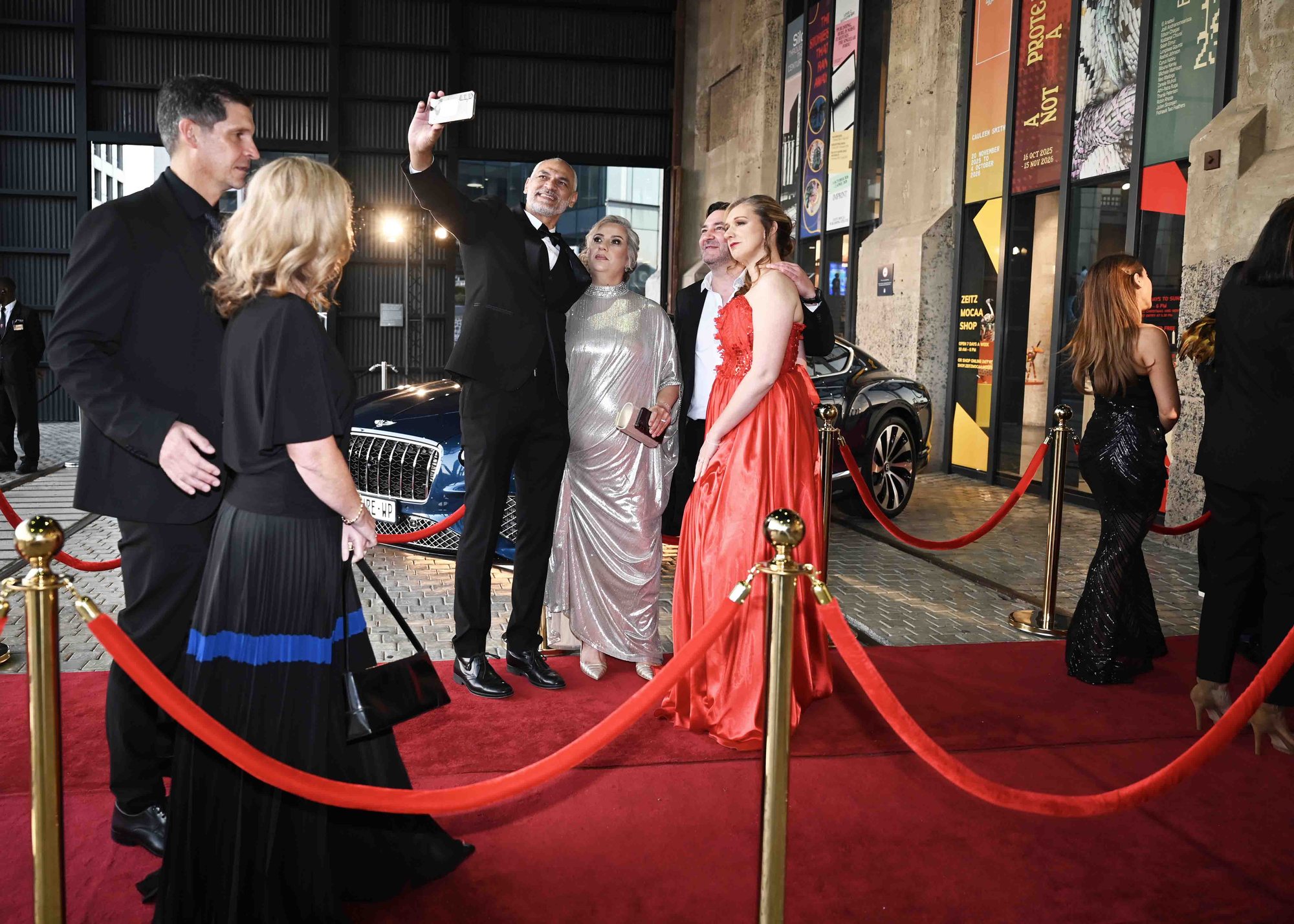 Guests in evening dress on the red carpet outside Zeitz MOCAA, one taking a selfie, with brass stanchions and red rope in the foreground.