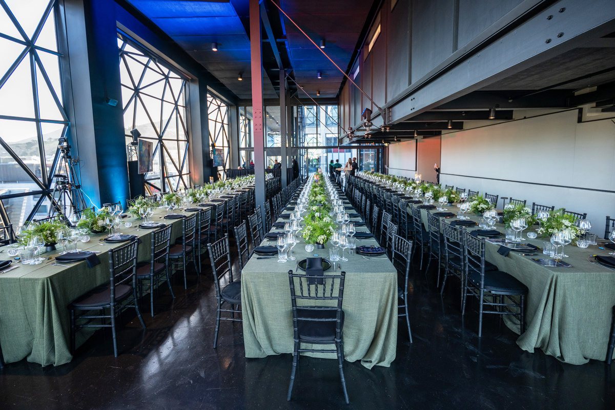 Long dining tables dressed in sage linen with low greenery centrepieces, running the length of a glass-walled hall at Zeitz MOCAA.