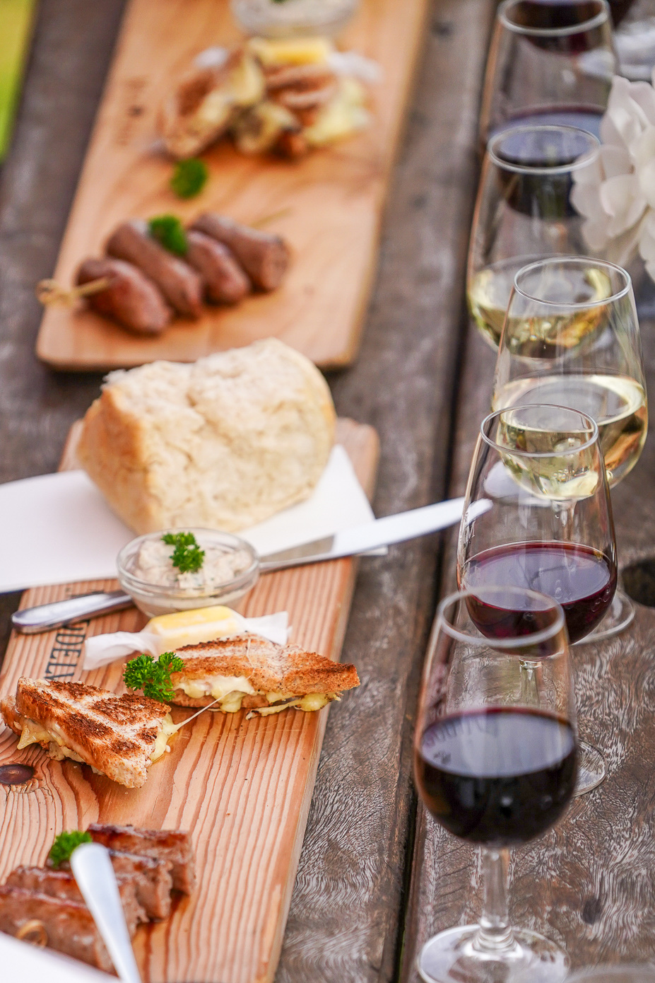 A wooden board of braaied boerewors, braaibroodjies and snacks beside a row of glasses of red and white wine