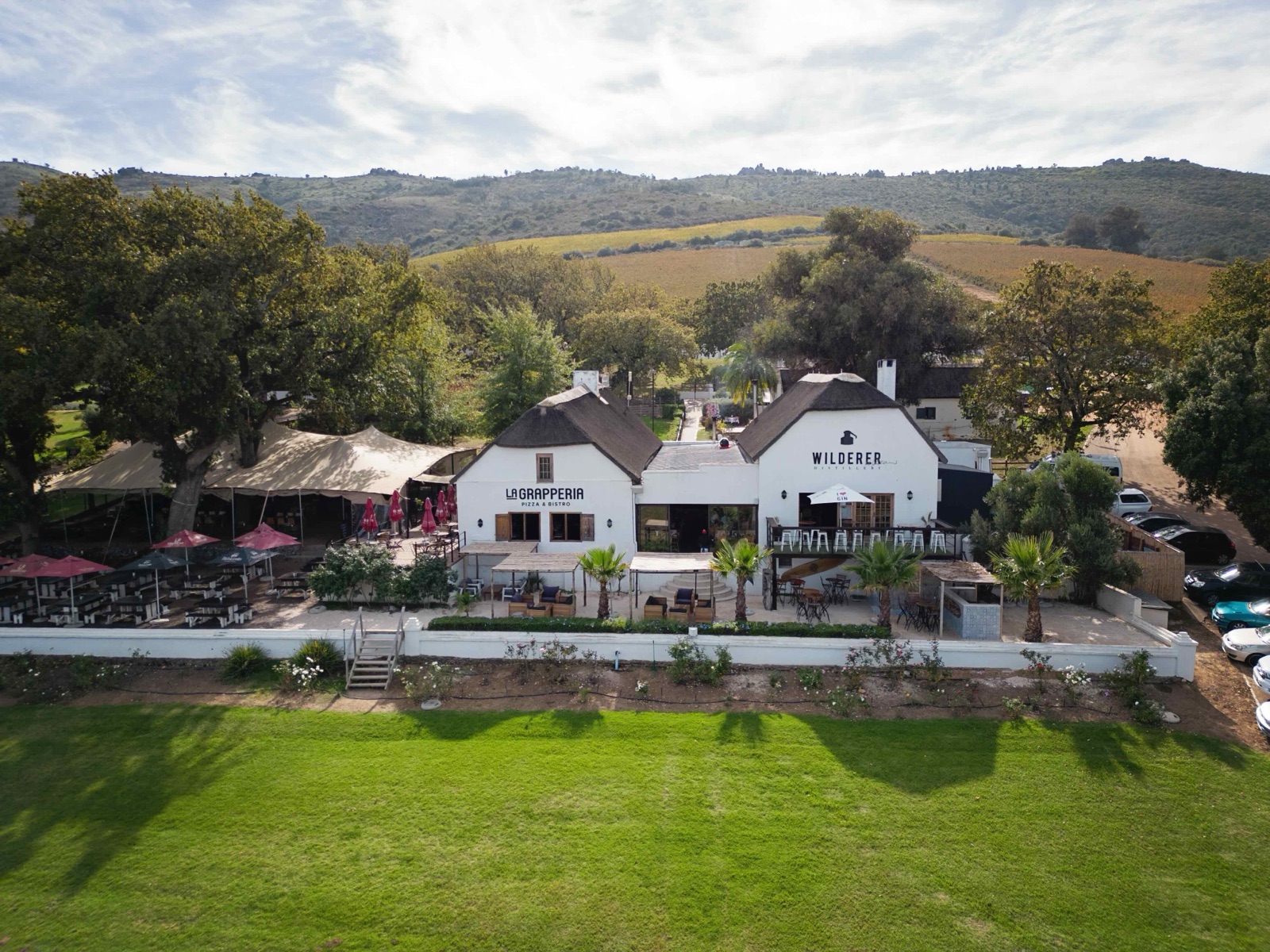 Aerial view of the Spice Route buildings, including La Grapperia, with umbrella-shaded terraces, a wide lawn and vineyards on the slopes behind