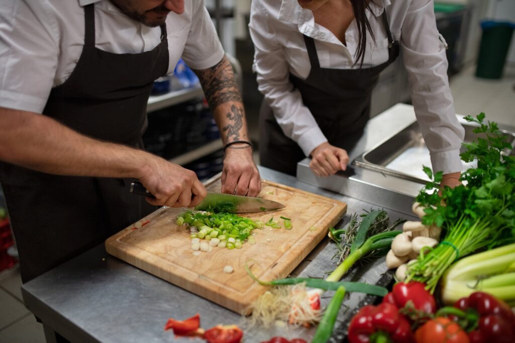 Two chefs in aprons chopping spring onions at a steel bench beside fresh vegetables